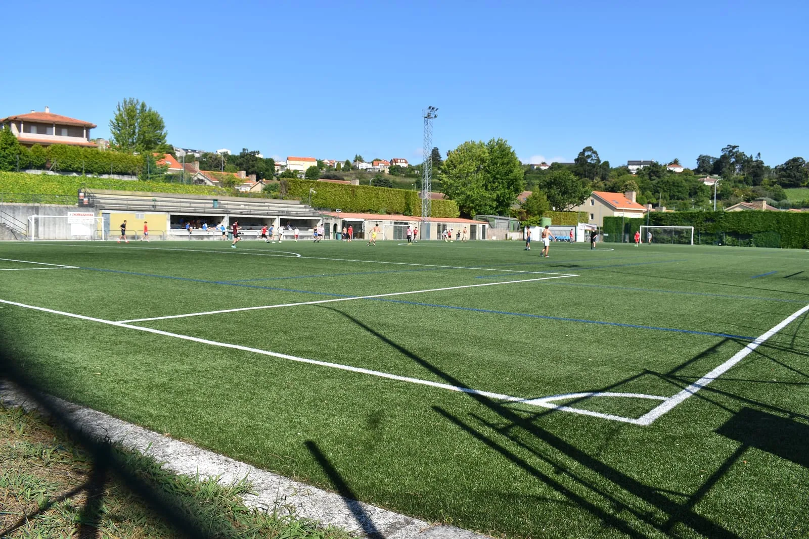 Vista general del campo municipal de O Redondo (Montrove, Oleiros): césped artificial con las líneas blancas y azules, edificio de vestuarios y torre de iluminación al fondo, casas y arboleda del entorno, y jugadores entrenando en la mitad lejana bajo un cielo despejado